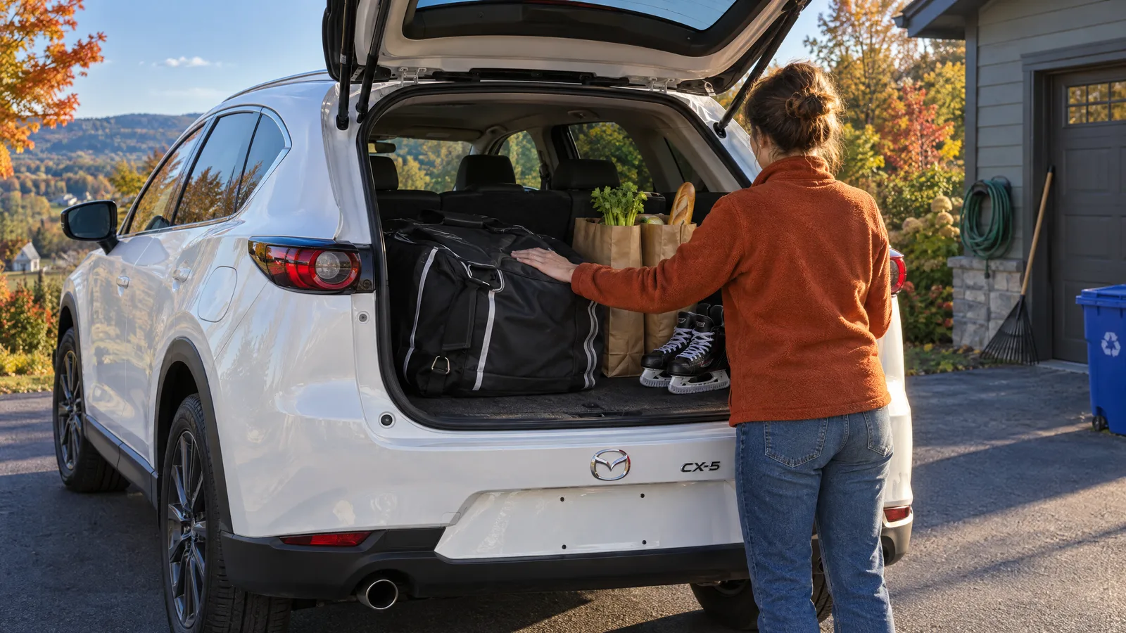 Personne de dos qui charge des sacs d'épicerie et un sac de hockey dans le coffre ouvert d'un Mazda CX-5 2026 blanc, entrée de cour en septembre