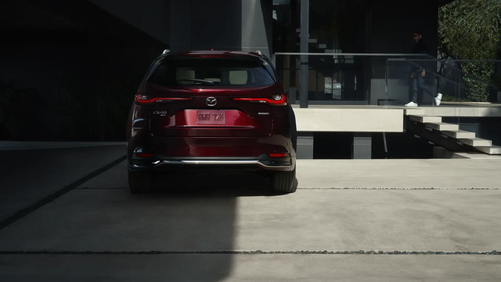 Mazda CX-90 2026 rouge vu de l'arrière, plaque et emblème visibles, stationné devant une résidence contemporaine
