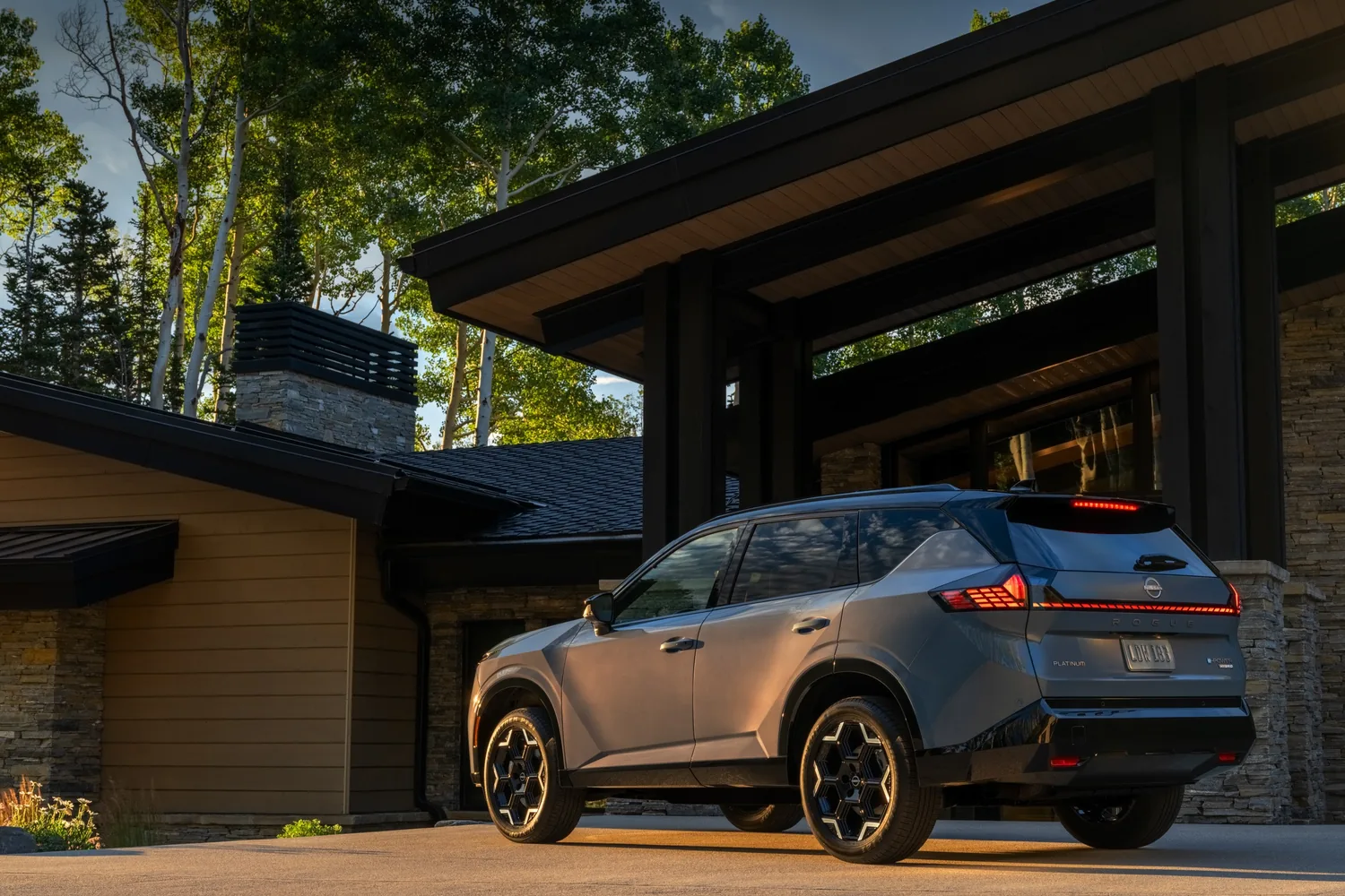 Nissan Rogue hybride 2027 gris vu de trois quarts arrière, stationné devant une maison moderne en bois et en pierre