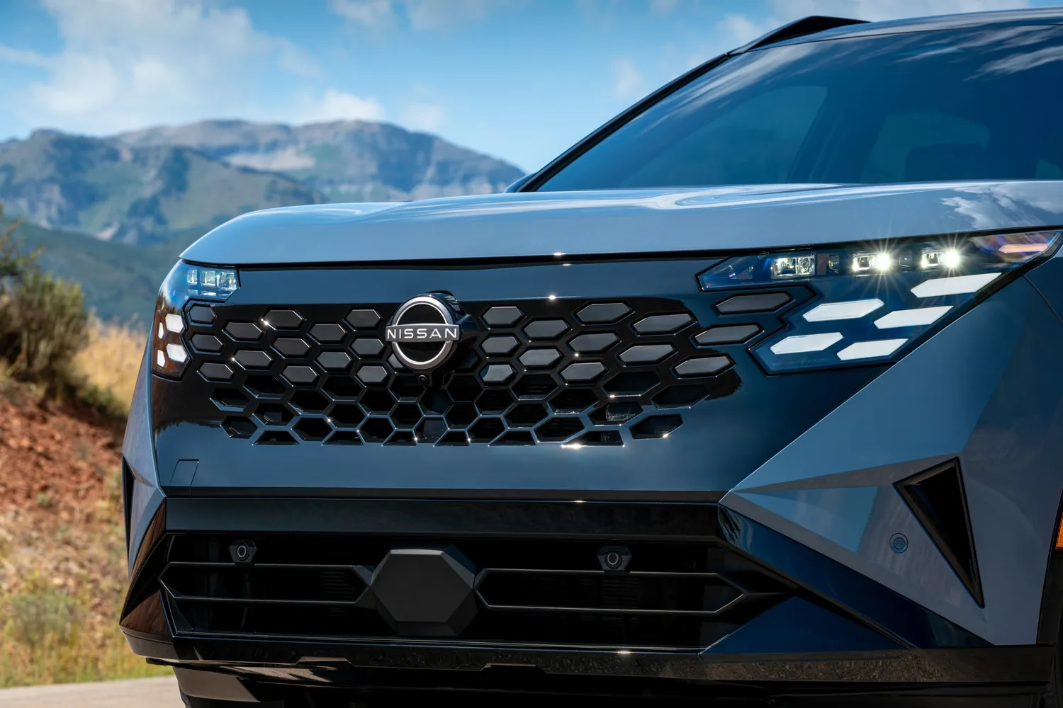 Gros plan sur la calandre à motif d'écailles et l'emblème Nissan éclairé d'un Rogue hybride 2027 bleu