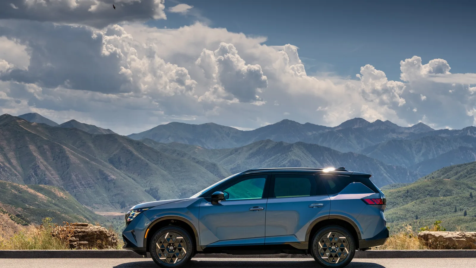 Nissan Rogue hybride 2027 bleu-gris de profil sur une route en lacets, chaîne de montagnes et ciel nuageux derrière