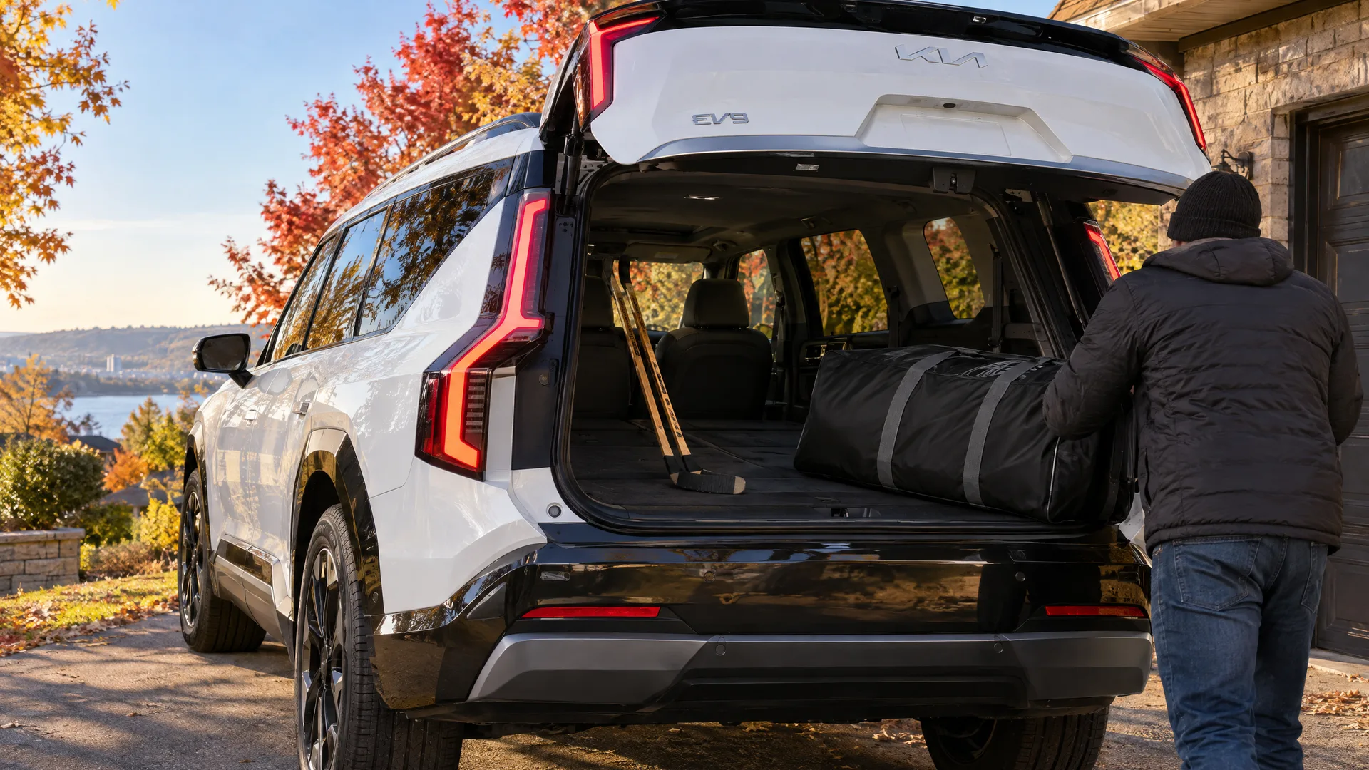 Personne vue de dos qui charge un sac de hockey dans le coffre d'un Kia EV9 2026 blanc, troisième rangée rabattue, entrée résidentielle québécoise en fin de journée