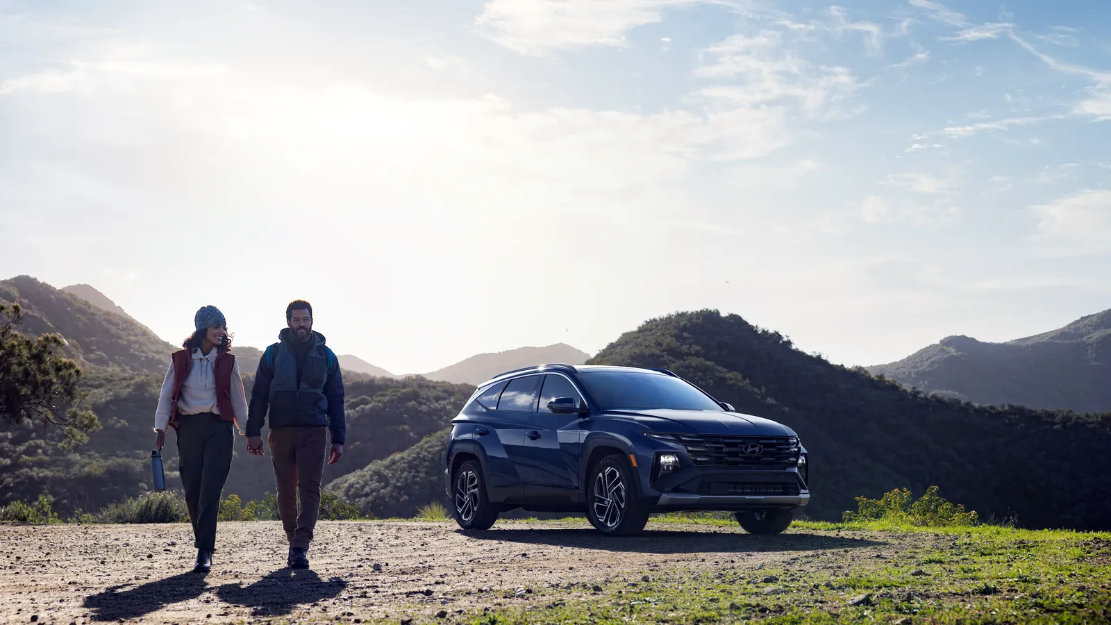Hyundai Tucson 2026 bleu foncé stationné sur un belvédère de montagne, un couple de randonneurs marchant au premier plan.