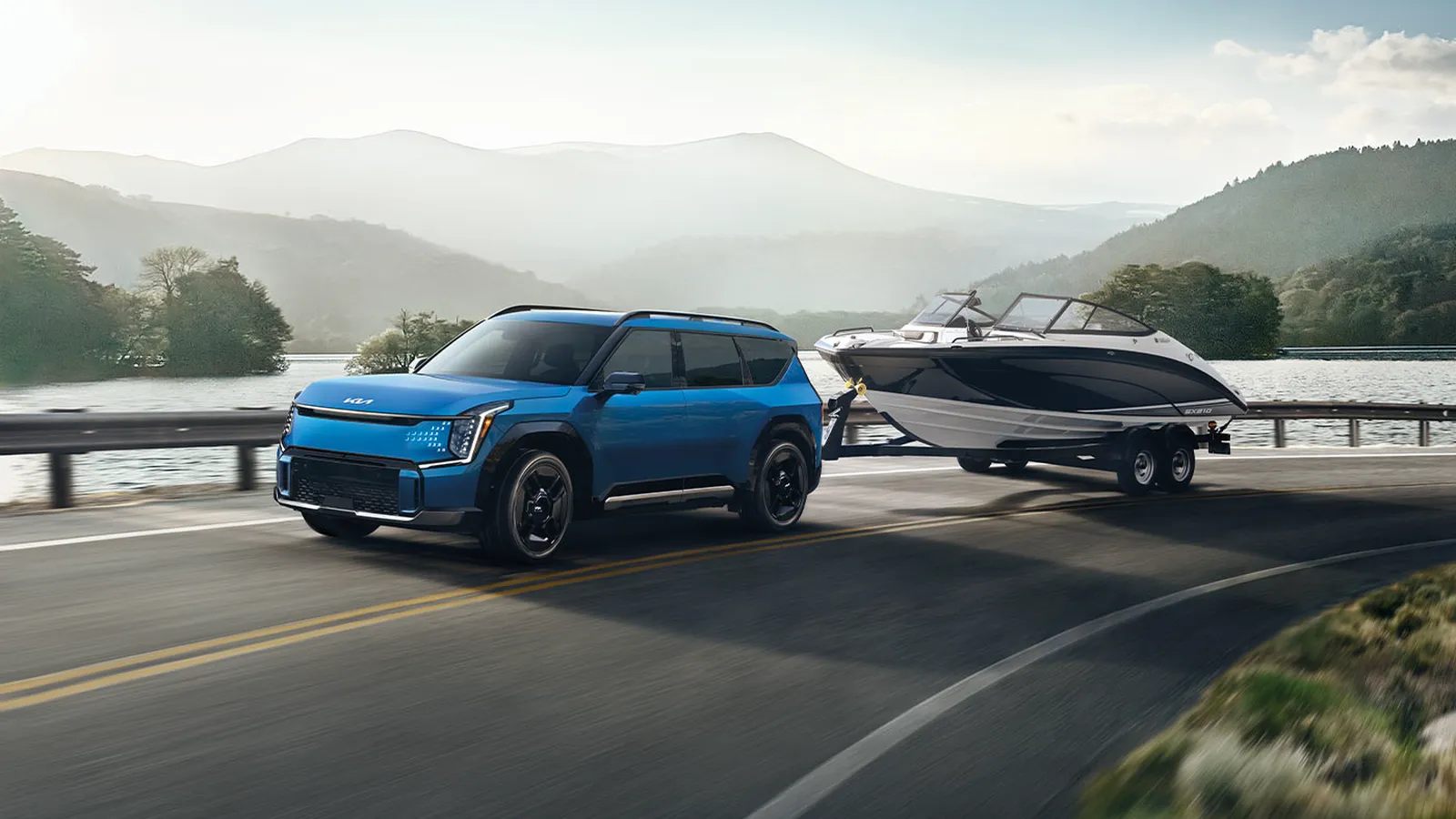 Kia EV9 2026 bleu tirant une remorque avec bateau sur une route de bord de lac entourée de montagnes, vue de trois quarts avant.