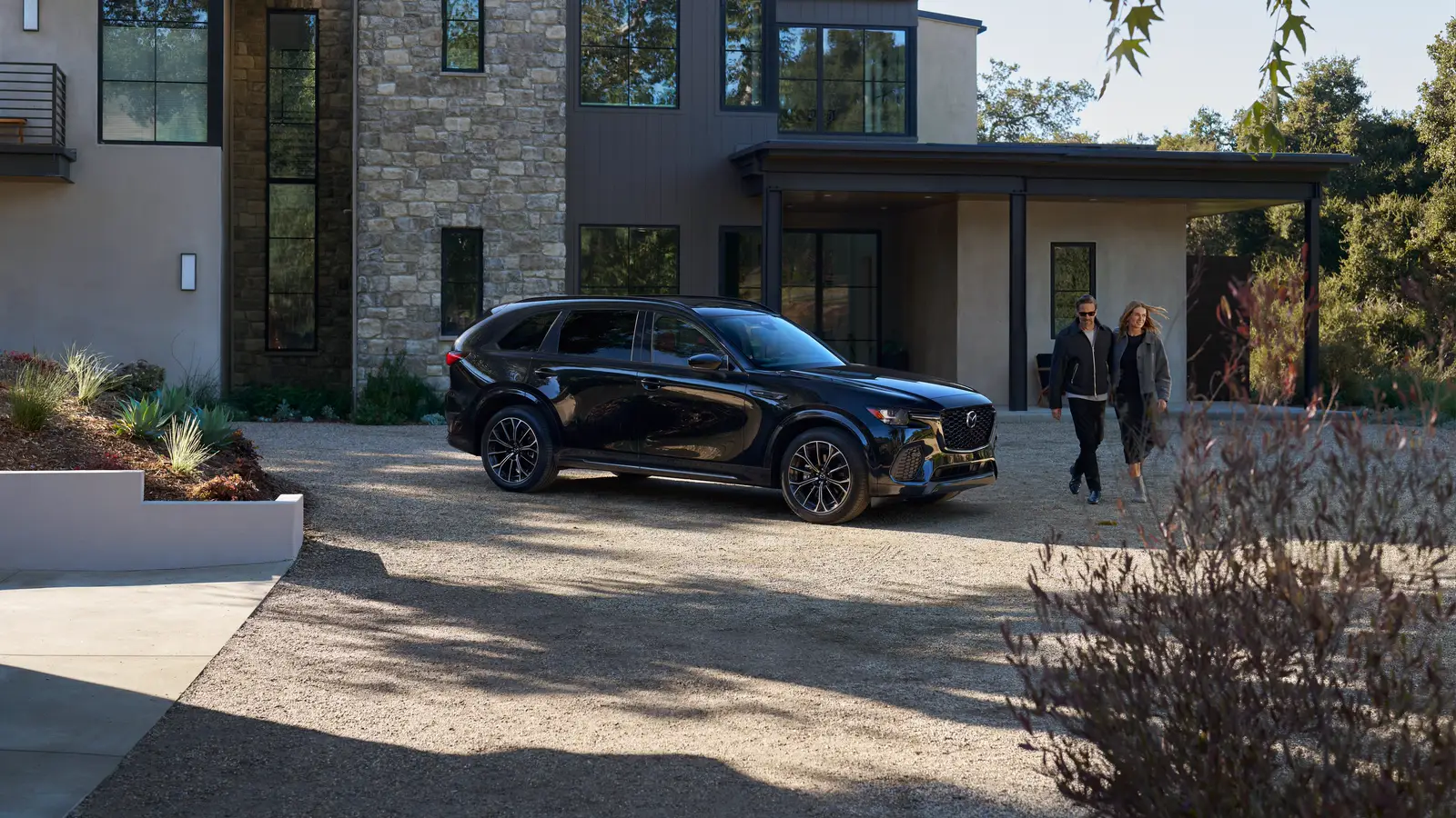 Mazda CX-70 2025 noire stationnée dans l’entrée gravillonnée d’une maison contemporaine en pierre, deux personnes marchant au loin.