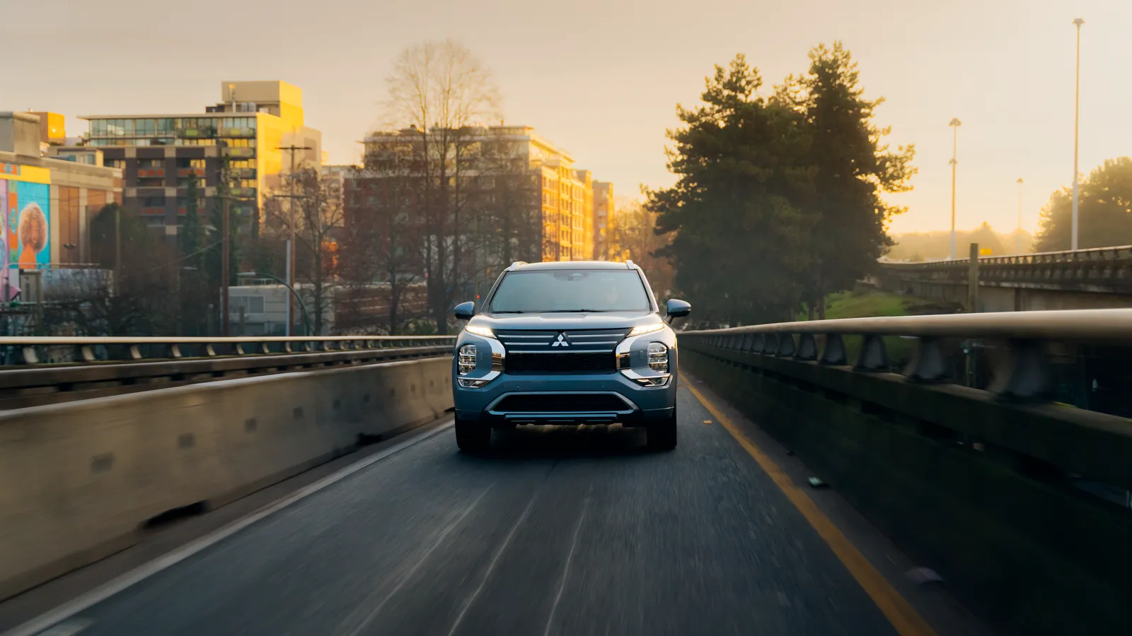 Mitsubishi Outlander PHEV 2026 bleu roulant sur une autoroute urbaine, silhouette de la ville en arrière-plan au coucher du soleil.