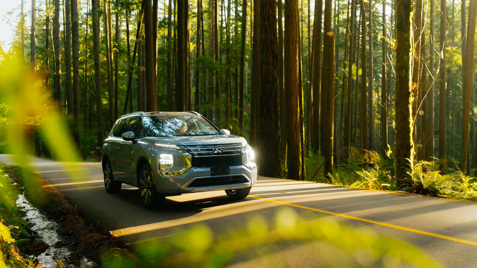 Mitsubishi Outlander PHEV 2026 vert roulant sur une route forestière bordée de grands conifères, lumière filtrée par les arbres.