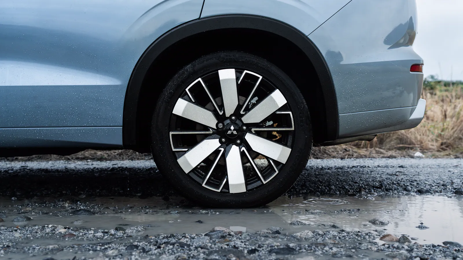 Gros plan sur la jante d’un Mitsubishi Outlander PHEV 2026 gris, emblème au centre du moyeu, chaussée de gravier mouillée.