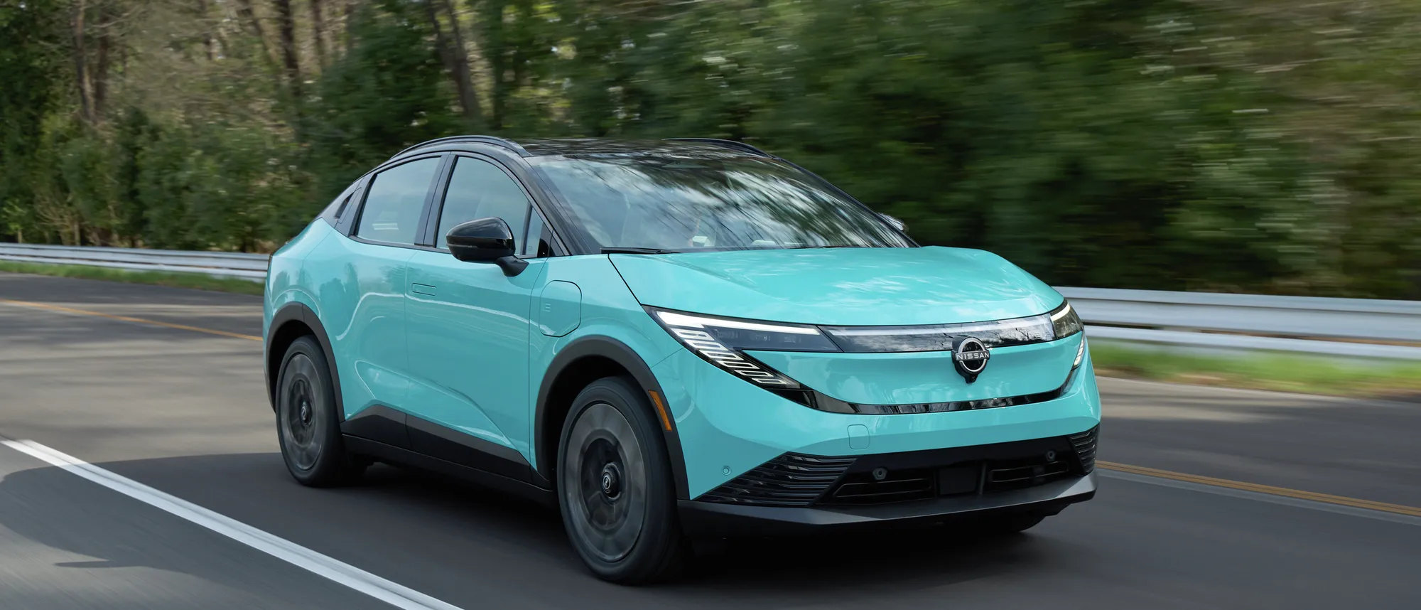 Nissan LEAF 2026 turquoise en roulage sur une route bordée de conifères, vue de trois quarts avant.