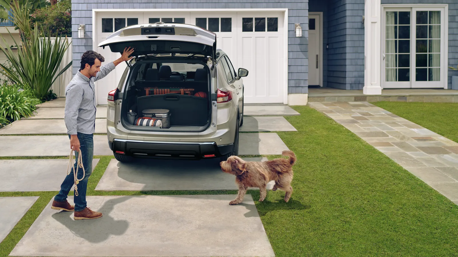 Un homme charge le coffre ouvert d’un Nissan Rogue 2027 gris stationné dans une entrée résidentielle, un chien à ses côtés.