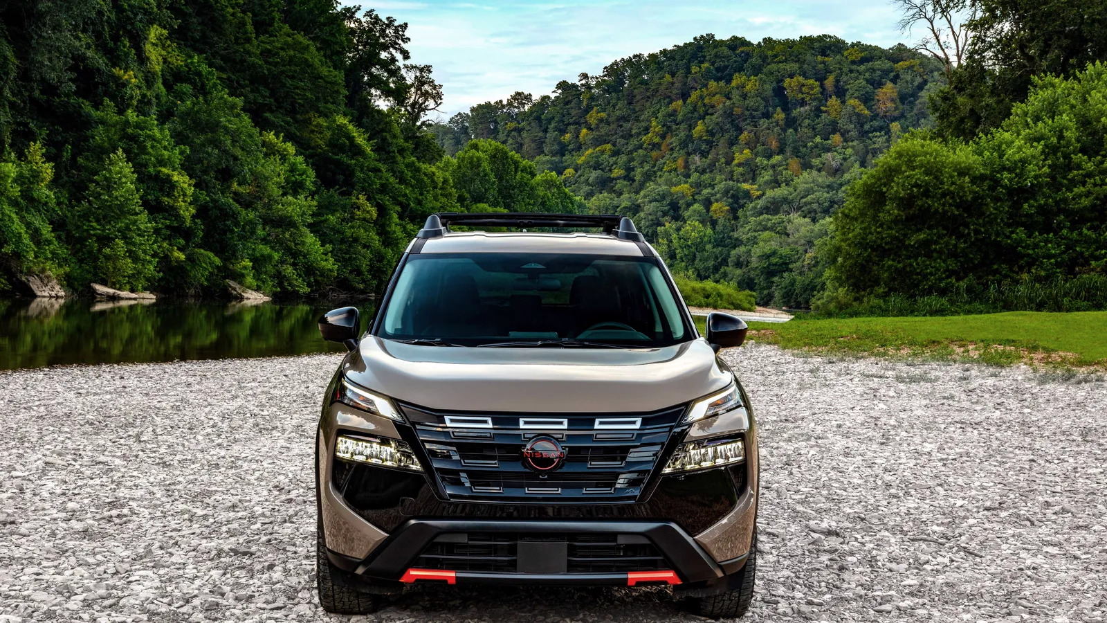 Nissan Rogue 2027 brun vue de face sur un chemin de gravier, entourée de montagnes et d’un cours d’eau.